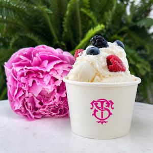 May include: A white paper cup filled with vanilla ice cream topped with fresh raspberries, blueberries, and a blackberry. The cup features a pink logo with the letters ST intertwined. A large, vibrant pink peony flower sits beside the cup.