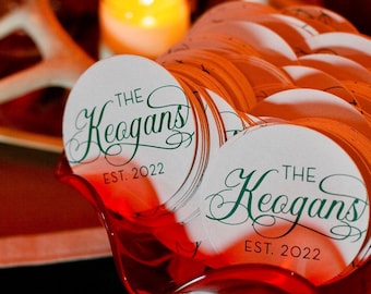 May include: A stack of circular white paper coasters featuring green cursive text that reads The Keogans and the text EST. 2022 in a simple sans-serif font. The coasters are contained within a translucent red scalloped glass bowl.