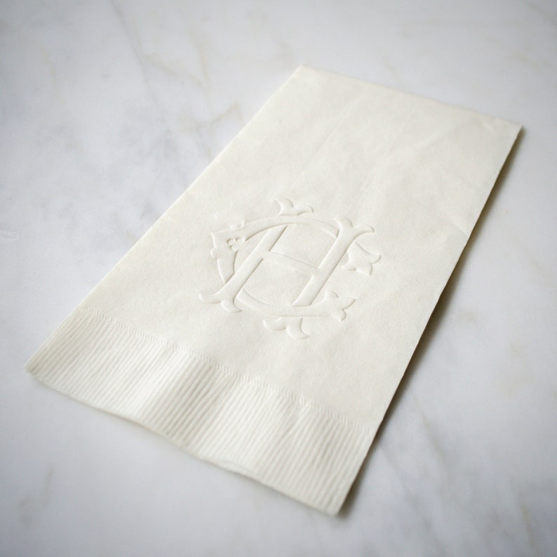 May include: A white linen napkin with an embossed monogram of the letter "H" in a decorative font.