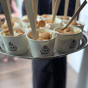 May include: A silver serving tray holds multiple white paper cups filled with banana pudding, each topped with a round vanilla wafer and a wooden spoon. The cups are printed with the black text Lisa's Bakery Est. 1965 and a small logo.