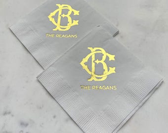 May include: Two square light gray paper napkins featuring a metallic gold foil monogram of the letters B and C intertwined. Below the monogram, the text THE REAGANS is printed in a matching metallic gold foil font on each of the napkins.