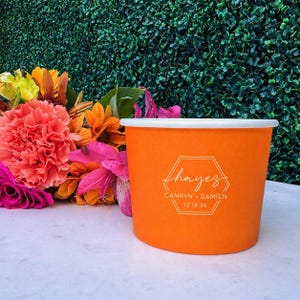May include: An orange paper cup featuring a white hexagonal graphic with the text "hayes", "CAMRYN + DAMIEN", and "12-18-26" printed on the front. The cup is positioned next to a vibrant arrangement of orange, pink, and yellow flowers.