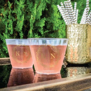 May include: Two clear plastic cups filled with a pink liquid, each featuring a gold-colored wreath design with the letters K and N printed in the center. A gold-colored hammered metal container holds several white and gold chevron-patterned straws.