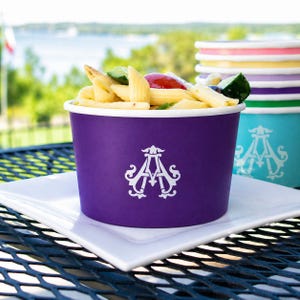 May include: A purple paper bowl filled with penne pasta salad containing cucumber and tomato pieces. The bowl features a white monogram logo with the letters AA. A stack of colorful paper bowls sits nearby, and the bowl rests on a white plate.