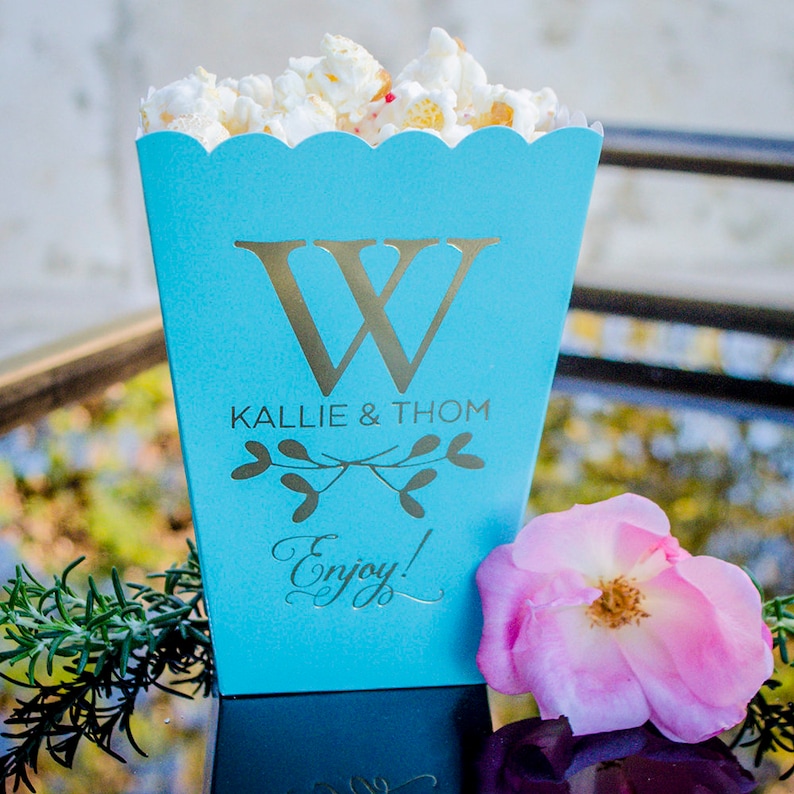 K&ouml;nnte beinhalten: Eine t&uuml;rkisfarbene Popcornbox mit einem goldenen Buchstaben "W" und den Namen "Kallie & Thom". Die Box ist mit Popcorn gef&uuml;llt und mit einer rosa Rose und Gr&uuml;nzeug verziert. Das Wort "Enjoy!" ist auf der Box aufgedruckt.