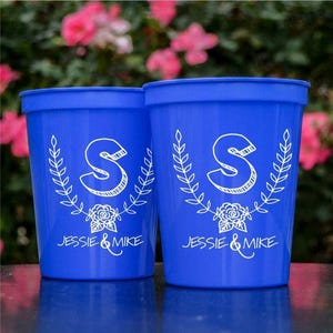 Könnte beinhalten: Zwei leuchtend blaue Stadionbecher aus Kunststoff mit weißem Grafikdruck. Das Design zeigt einen großen Buchstaben S über einem Blumenkranz und den Text Jessie & Mike. Die Becher haben eine glatte Oberfläche und einen breiten, offenen Rand.