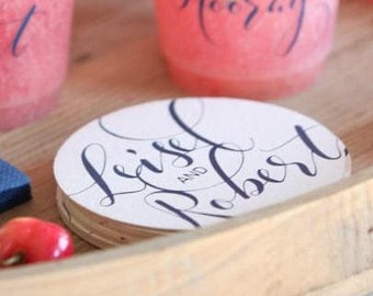 May include: A stack of round white paper coasters featuring dark blue cursive calligraphy that reads Leisel and Robert. The coasters are positioned on a wooden surface near a red cherry and a pink cup with the text Hooray.