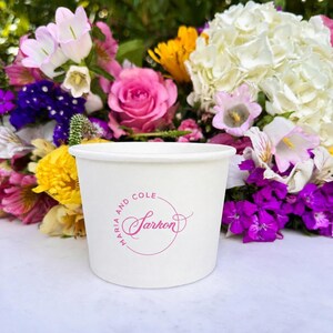 May include: A white paper cup featuring a pink circular logo with the text Maria and Cole and the name Sarkon in a decorative script font. The cup is positioned in front of a vibrant arrangement of pink, yellow, white, and purple flowers.