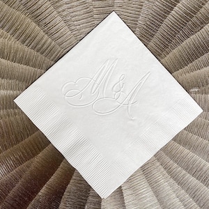 May include: A square white paper napkin featuring an embossed monogram of the letters M and A joined by an ampersand in the center. The napkin has a textured border along its edges, creating a subtle decorative frame around the central design.