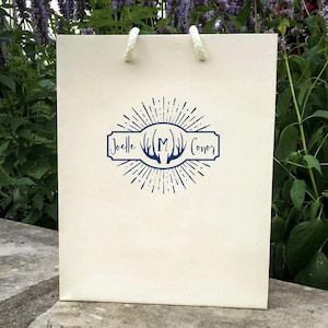 May include: A white gift bag with a blue monogram design. The monogram features a stylized antler design with the letter "M" in the center. The text "Joelle" and "Conor" are on either side of the antler design.
