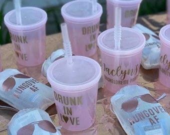 May include: Light pink plastic cups with lids and straws, featuring gold text that reads "Drunk in Love" or "Jaclyn's Bachelorette Party." Accompanying the cups are small white drawstring bags printed with sunglasses and the text "Hungover AF."
