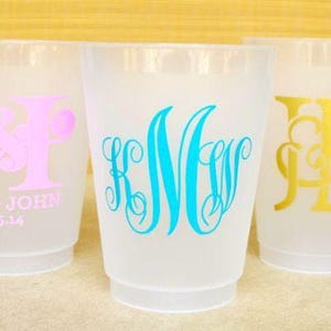 May include: Three frosted plastic cups featuring custom monograms and text. The left cup has a pink monogram with the text "ERIN JOHN 4-5-14". The center cup displays a bright blue monogram, and the right cup features a gold monogram with an ampersand.
