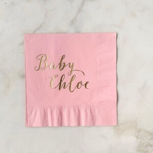 May include: A square, light pink paper napkin featuring the text Baby Chloe printed in a metallic gold cursive script. The napkin has a textured, ribbed border along the bottom edge and a smooth finish on the main surface area.