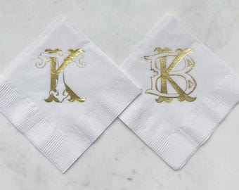 May include: Two square white paper napkins featuring decorative gold foil monograms. The left napkin displays a stylized letter K, while the right napkin features a stylized letter B. Both napkins have a textured border and a smooth center.