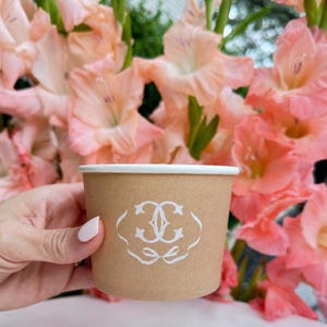May include: A light brown paper cup with a white rim and a white decorative emblem printed on the front. The emblem features a stylized double C monogram enclosed within a delicate, flowing line design that resembles a ribbon or bow.
