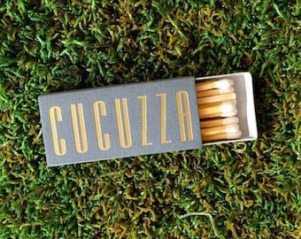 Puede incluir: Una caja de cerillas rectangular de color gris con la palabra CUCUZZA impresa en letras mayúsculas doradas. La caja está parcialmente abierta, revelando una fila de cerillas de madera con puntas claras dispuestas ordenadamente en la bandeja.