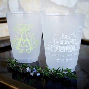 May include: Two frosted plastic cups. One cup features a large, ornate, light green letter A. The other cup displays a white line drawing of a house with the text MICHELLE & JONATHAN, CHARLESTON, SC, APRIL 6, 2018 printed below the illustration.