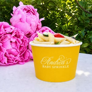May include: A yellow paper bowl filled with penne pasta, cucumber slices, and cherry tomatoes. The bowl features white text that reads Andrea's BABY SPRINKLE. The bowl is positioned in front of large, vibrant pink peony flowers.