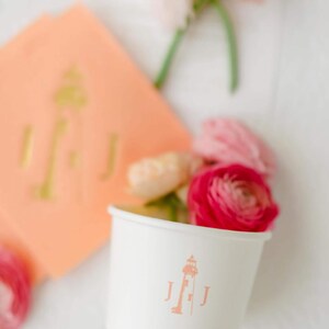May include: A white paper cup featuring a peach-colored lighthouse graphic flanked by the letter J on both sides. The cup contains pink and cream-colored flowers, and a matching peach-colored napkin with a gold lighthouse monogram is nearby.