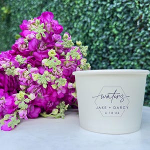 May include: A white paper cup featuring a black printed hexagon design with the text "waters", "JAKE + DARCY", and "6.18.26" positioned next to a cluster of vibrant magenta flowers with small green buds and stems.