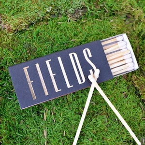 May include: A rectangular black matchbox with the word FIELDS printed in a tall, white, sans-serif font. The box is partially open, revealing a row of wooden matches with white tips. Two additional matches are crossed in front of the box.