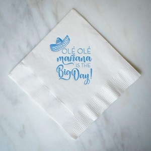 May include: A square white paper napkin featuring a light blue printed graphic of a sombrero and the text "OLÉ OLÉ mañana IS THE Big Day!" in a mix of block and script fonts. The napkin has a textured, embossed border along its four edges.
