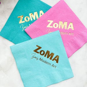 Könnte beinhalten: Drei quadratische Papierservietten in Türkis, Pink und Hellblau. Jede Serviette zeigt den goldfarbenen Foliendruck "ZoMA" in einer fetten serifenlosen Schrift sowie den Schriftzug "Zoe's Modern Art" in kleinerer Schreibschrift darunter.