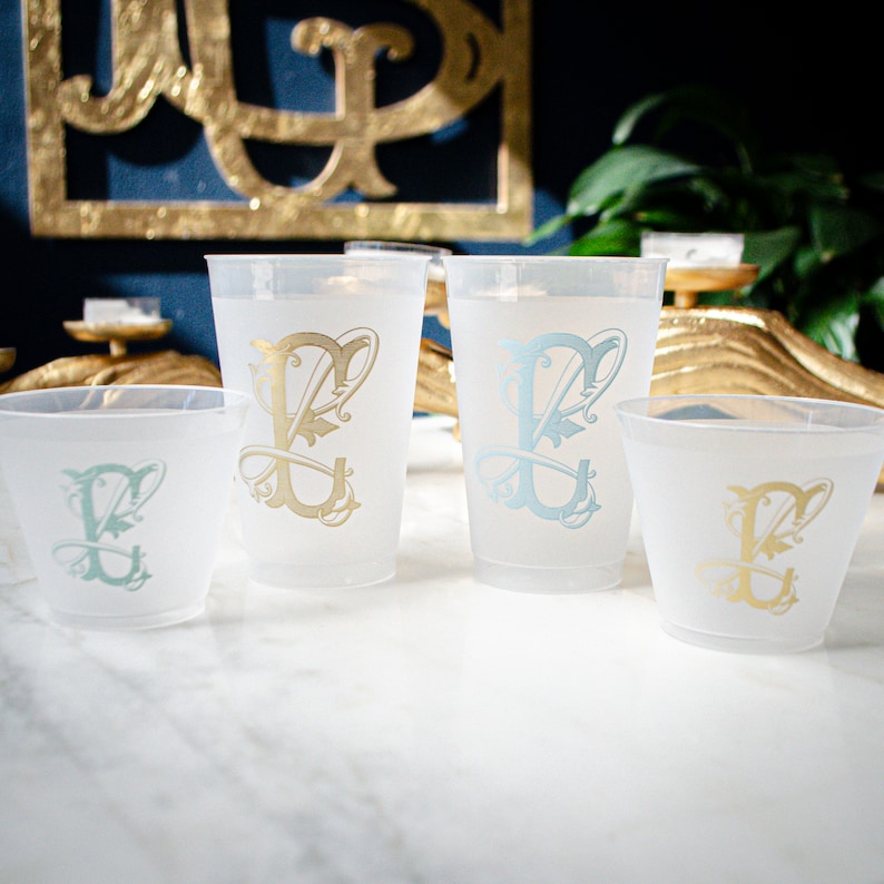 May include: Four frosted plastic cups with a gold and blue monogram design. The cups are arranged in a row on a white marble surface.