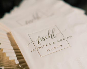 May include: A stack of white paper napkins featuring a gold foil-stamped design. The print includes a rectangular border, the name fischl in script, and the text JENNIFER & ADRIAN 11.16.19 printed in a clean, sans-serif capital typeface.