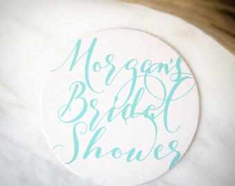 May include: A round, white coaster featuring the text "Morgan's Bridal Shower" written in a decorative, flowing light blue script font. The coaster has a smooth, matte surface and is positioned flat, displaying the centered lettering clearly.