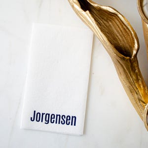 May include: A rectangular white paper napkin featuring the name Jorgensen printed in a dark blue sans-serif font. The napkin is positioned next to a textured gold-colored decorative object with a curved, organic shape and a metallic finish.