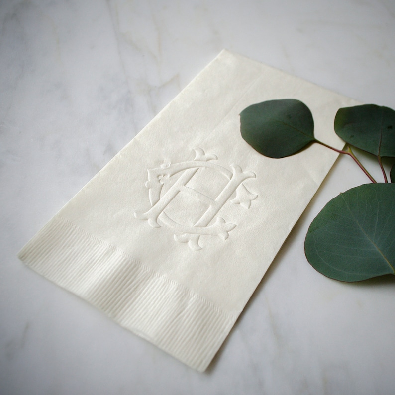 May include: A white linen napkin with an embossed monogram of the letter "H" surrounded by a decorative design. The napkin is on a white marble surface with a sprig of green eucalyptus leaves.