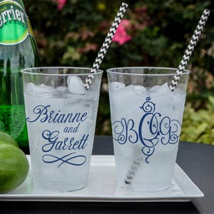 Könnte beinhalten: Zwei durchsichtige Kunststoffbecher gefüllt mit Eis und Flüssigkeit, jeweils mit dunkelblauem Text versehen. Ein Becher zeigt die Namen Brianne und Garrett in Schreibschrift, der andere ein dekoratives Monogramm mit den Buchstaben B, C und G. In beiden Bechern stecken schwarz-weiß gemusterte Papierstrohhalme.