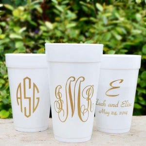 May include: Three white foam cups featuring gold lettering. The left cup displays a three-letter monogram. The center cup features a large, ornate script monogram. The right cup shows a script letter E above the text Zach and Elise May 24, 2014.