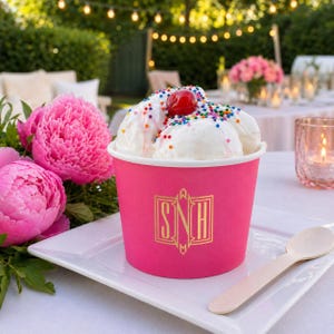May include: A bright pink paper cup filled with scoops of white ice cream topped with colorful sprinkles and a single red cherry. The cup features a gold monogram with the letters SNH printed on the front. A wooden spoon rests on the white plate.
