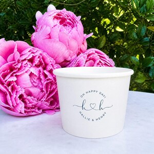 May include: A white paper cup featuring a circular black logo with the text OH HAPPY DAY! and HALLIE & HENRY surrounding a stylized H and h connected by a heart. The cup is positioned in front of three large, vibrant pink peony flowers.