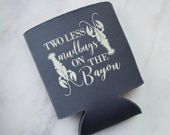 May include: A dark gray foam can cooler featuring the white printed text Two Less mudbugs ON THE Bayou, accompanied by two white illustrations of crawfish positioned on the left and right sides of the lettering.