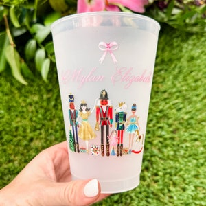 May include: A white plastic cup with a frosted finish features a pink ribbon and the name "Mylan Elizabeth" written in pink script. The cup has a colorful illustration of characters from the Nutcracker ballet, including the Nutcracker, Clara, and the Mouse King.