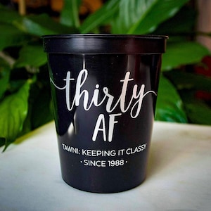 May include: A black plastic stadium cup featuring white text that reads thirty AF in a script font, followed by the text TAWNI: KEEPING IT CLASSY SINCE 1988 in a clean sans-serif font. The cup has a smooth finish and a wide, circular rim.