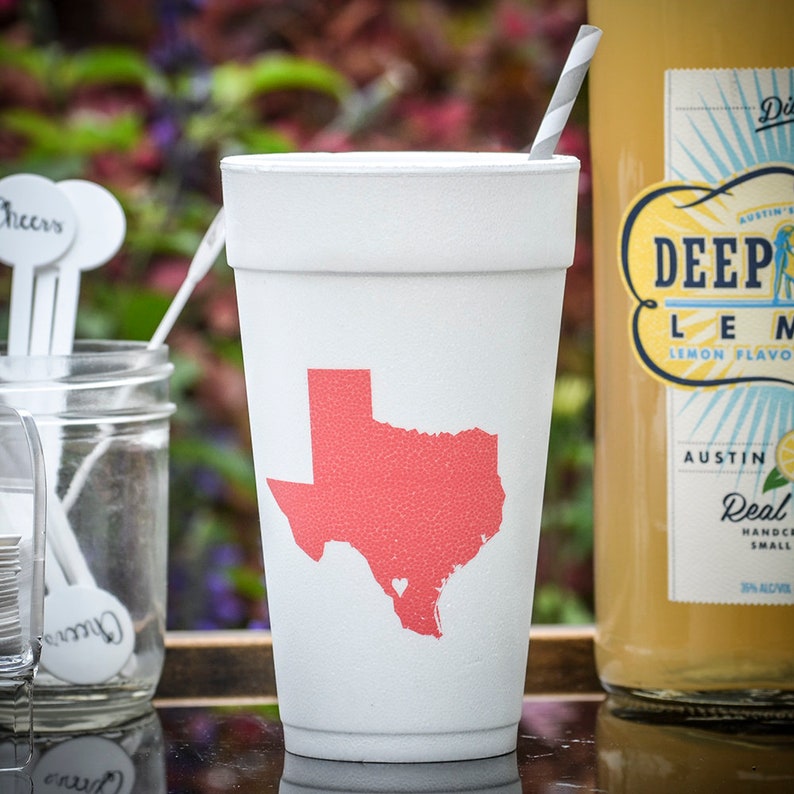 May include: White foam cup with a pink outline of the state of Texas with a small pink heart in the center. The cup is sitting on a wooden surface next to a bottle of Deep Eddy Lemon flavored vodka.