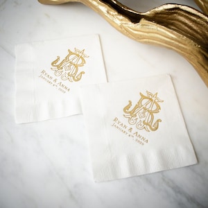 May include: Two square white paper napkins featuring a gold foil monogram of the letters R and A. Below the monogram, the text reads RYAN & ANNA and JANUARY 4TH, 2020 in gold lettering. The napkins have a textured border along the bottom edge.
