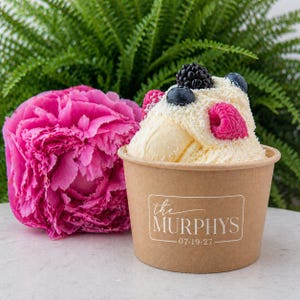 May include: A brown paper cup filled with vanilla ice cream topped with fresh blackberries, blueberries, and raspberries. The cup features a white rectangular label with the text the MURPHYS and the date 07-19-27 printed in a white serif font.