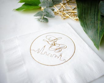 May include: A square white paper napkin featuring a gold foil stamped design. The design consists of a circular border enclosing a stylized city skyline silhouette at the bottom and the elegant cursive monogram letters L and B joined by an ampersand.
