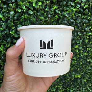 May include: A white paper cup featuring a black logo with the text LUXURY GROUP and MARRIOTT INTERNATIONAL printed on the front. The cup has a smooth, matte finish and a simple, cylindrical shape with a slightly wider rim at the top edge.