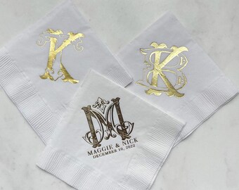 May include: Three white square paper napkins featuring metallic gold foil monograms. Two napkins display large individual letters, while the third features an ornate monogram with the text MAGGIE & NICK and the date DECEMBER 10, 2022 printed below.