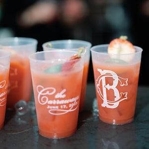 May include: Several translucent plastic cups filled with a pinkish-orange cocktail, each garnished with a strawberry slice. The cups feature white printed text that reads "the Carraways JUNE 17, 2023" and a large stylized letter "B" logo.
