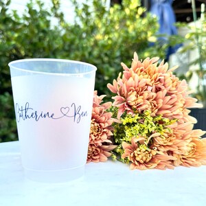 May include: A frosted plastic cup with the names "Catherine & Ben" in a cursive font, with a heart symbol between the names. A bouquet of peach-colored flowers is next to the cup. The background is blurred green foliage.