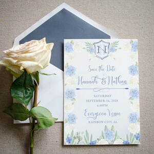 May include: Save the date card with a watercolor floral border in shades of blue and white. The card features the initials "H & N" in a crest and the text "Save the Date Hannah & Nathan Saturday September 16, 2028 6:00PM Evergreen Venne RAINBOW CITY, AL"