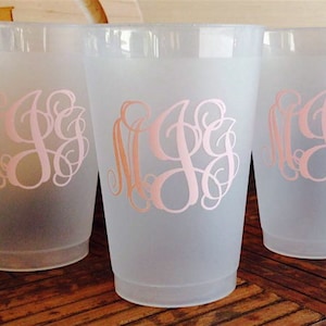 May include: Three frosted plastic cups with pink monogrammed designs. The cups are clear and have a frosted finish. The monograms are in a script font and are centered on the front of each cup.