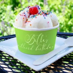 May include: A lime green paper cup filled with white ice cream scoops topped with colorful sprinkles and a single red cherry. The cup features the white cursive text Bella's third birthday. A wooden spoon rests on the white square plate.